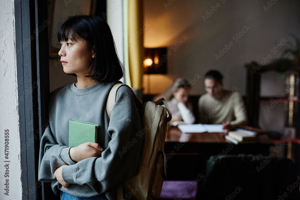 Pensive young woman with books looking through the window while standing in the room Pensive young woman with books looking through the window while standing in the room
