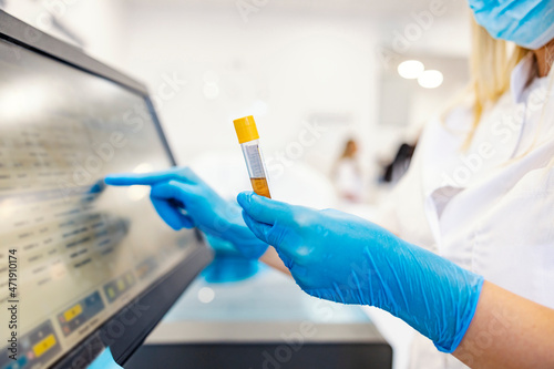 A nurse with a mask on her face and with sterile gloves is holding a test tube with a blood sample and entering test results into the computer. The nurse measured corona antibodies in a blood sample.