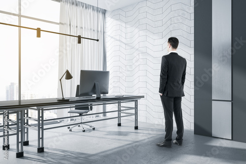 Attractive businessman standing in modern concrete office interior with window and city view, daylight, furniture and various objects. Workplace concept.