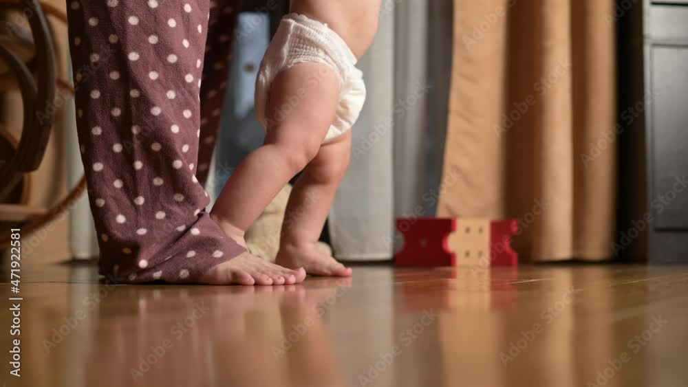 The first steps of a little boy around the house. The baby in the diaper learns to walk with the help of the mother. Close-up of legs