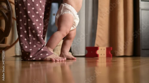 The first steps of a little boy around the house. The baby in the diaper learns to walk with the help of the mother. Close-up of legs