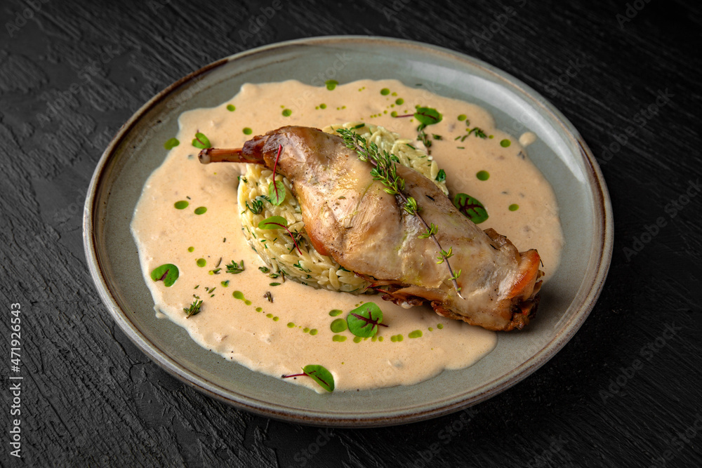 Rabbit leg baked with rice in sauce in a ceramic plate on a dark textured background. Restaurant menu Isolated on black