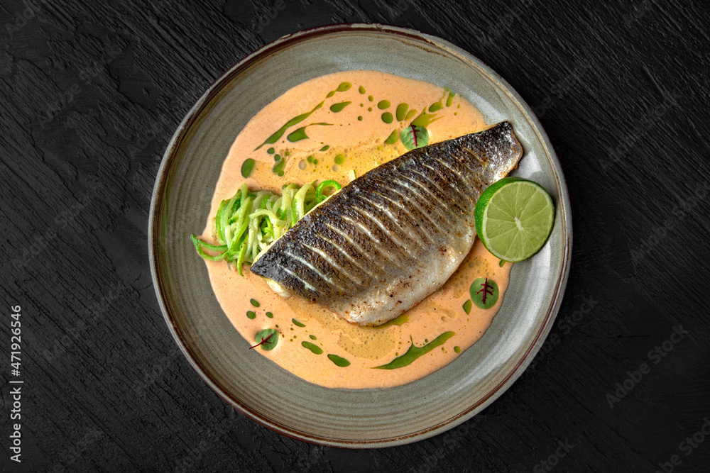 Seabass fish baked with tender sauce on seafood broth in a ceramic plate on a dark textured background. Restaurant menu Isolated on black Seabass fish baked with tender sauce on seafood broth in a ceramic plate on a dark textured background. Restaurant menu Isolated on black