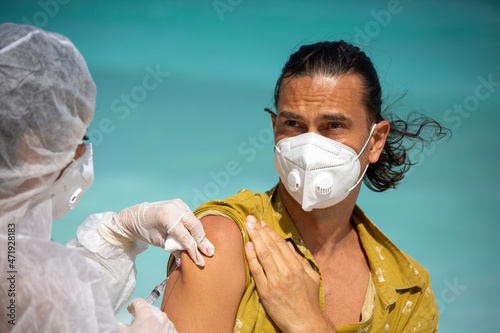 A doctor in a protective suit gives a covid-19 vaccine to a young attractive European man in a yellow shirt with long hair on a tropical beach. Against the background of the turquoise sea