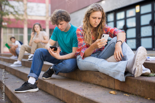 Teenager girl sitting on stairs beside school building and using smartphone. Her classmate sitting next to her and using smartphone too.