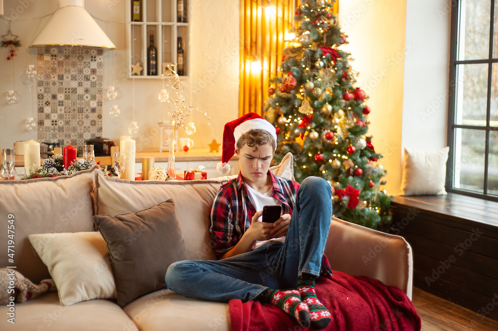 Teenage boy sits on the couch wearing a santa hat and with a phone in his hands. Online communication at Christmas. Holiday online shopping. Christmas atmosphere and relaxation. Teenage boy sits on the couch wearing a santa hat and with a phone in his hands. Online communication at Christmas. Holiday online shopping. Christmas atmosphere and relaxation.