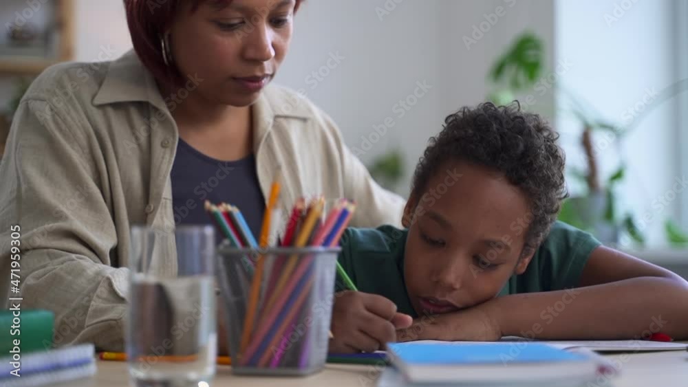 Distant home schooling of junior children. Young black mother cleans stained cheek and mouth of son student coloring notebook page at table in living room spbd Distant home schooling of junior children. Young black mother cleans stained cheek and mouth of son student coloring notebook page at table in living room spbd