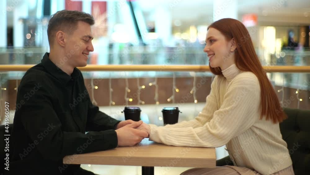 Side view of beautiful young couple drinking coffee, holding hands and talking looking at each other on date sitting at table in coffee shop. Cute redhead lady chatting joyfully with handsome male. Side view of beautiful young couple drinking coffee, holding hands and talking looking at each other on date sitting at table in coffee shop. Cute redhead lady chatting joyfully with handsome male.