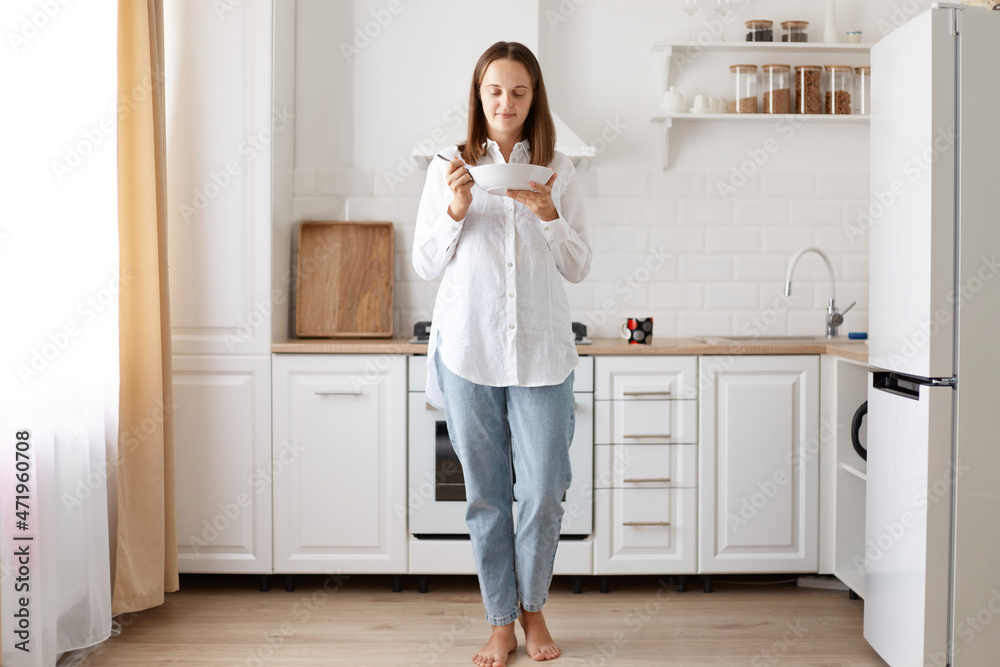 Portrait of happy young housewife eating soup, holding plate in hands while standing against while kitchen set, wearing jeans and shier, having breakfast before going to work.