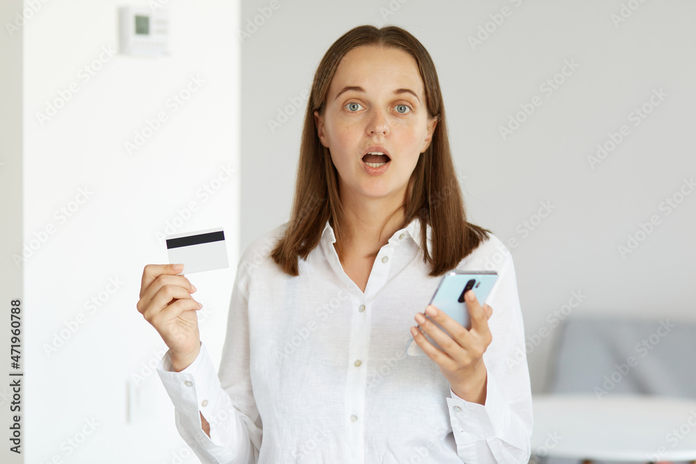 Shocked amazed woman wearing white shirt standing indoor in light room holding credit card and cell phone in hands, being surprised of winning lottery or betting. Shocked amazed woman wearing white shirt standing indoor in light room holding credit card and cell phone in hands, being surprised of winning lottery or betting.