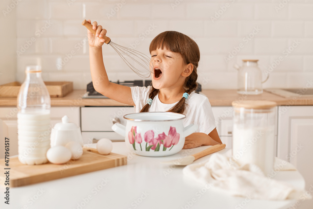 Indoor shot of excited little cute female kid with dark hair wearing white t shirt cooking in light kitchen, holding corolla in hands and singing like microphone while cooking. Indoor shot of excited little cute female kid with dark hair wearing white t shirt cooking in light kitchen, holding corolla in hands and singing like microphone while cooking.