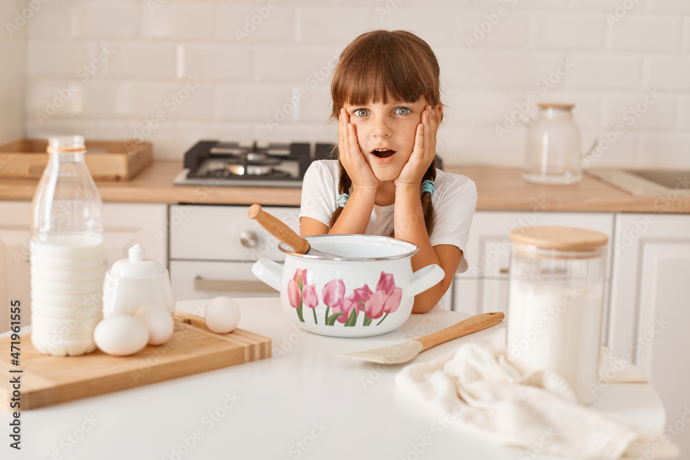 Portrait of surprised amazed little cute girl with dark hair and braids wearing white t shirt cooking in light kitchen, looking at camera with open mouth, forgot recipe of her favorite cakes. Portrait of surprised amazed little cute girl with dark hair and braids wearing white t shirt cooking in light kitchen, looking at camera with open mouth, forgot recipe of her favorite cakes.