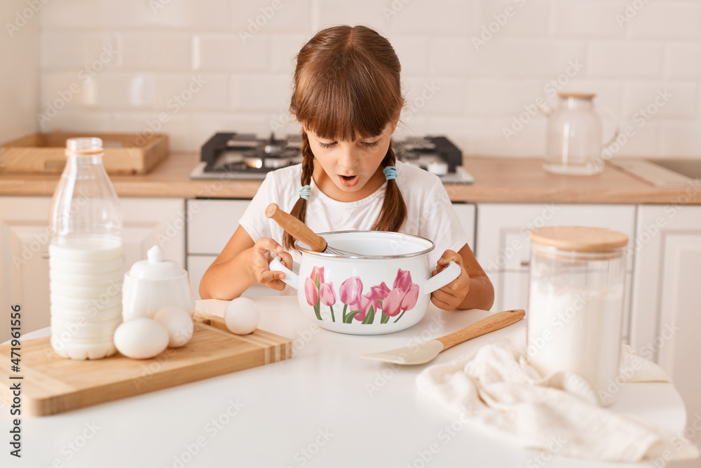 Indoor shot of shocked amazed little cute female child with dark hair wearing white t shirt baking in light kitchen, looking inside pot with shocked expression, making mistake, keeps mouth opened. Indoor shot of shocked amazed little cute female child with dark hair wearing white t shirt baking in light kitchen, looking inside pot with shocked expression, making mistake, keeps mouth opened.