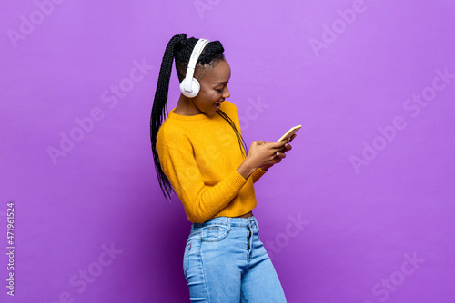 African American woman wearing headphones and listening to music from smart phone on colorful purple isolated studio background
