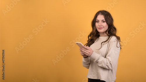 Attractive and happy Caucasian brunette girl with wavy hair in a casual jumper using a smartphone on an orange studio background with copy space. © Павел Костенко