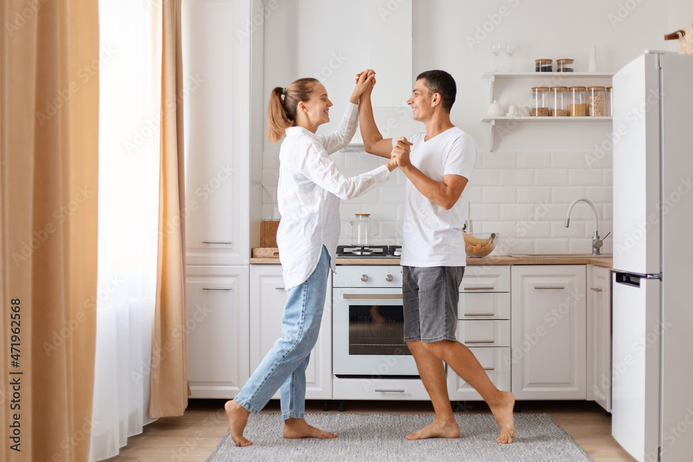 Fototapeta premium Indoor shot of happy beautiful couple smiling while dancing in kitchen at home, holding each other hands, enjoying spending time together, family wearing white shirts and jeans.