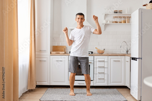 Carefree funny young adult brunette man having fun, dancing alone in modern kitchen interior, active happy single guy enjoying silly movements, listening music celebrating freedom.