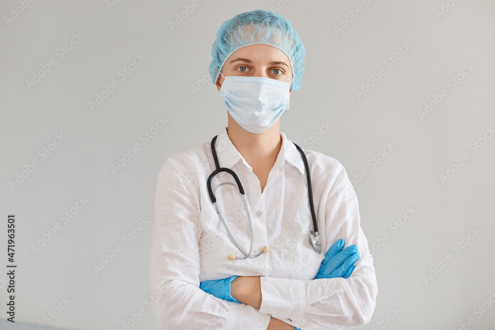 Indoor shot of young adult Caucasian doctor woman wearing gloves, medical mask and cap, gown, standing with folded hands and looking at camera, working in hospital or clinic. Indoor shot of young adult Caucasian doctor woman wearing gloves, medical mask and cap, gown, standing with folded hands and looking at camera, working in hospital or clinic.