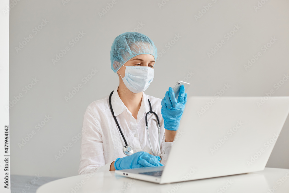 Confident woman doctor sitting at table and working on laptop computer, holding mobile phone in hands for checking e mails, wearing medical cap, surgical mask, rubber gloves and gown. Confident woman doctor sitting at table and working on laptop computer, holding mobile phone in hands for checking e mails, wearing medical cap, surgical mask, rubber gloves and gown.