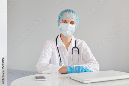 Indoor shot of serious confident female doctor wearing medical cap  surgical mask  rubber gloves and gown  using notebook for working  looking at camera while sitting at table.