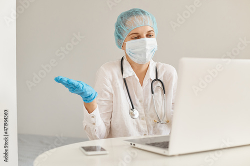 Confused female doctor wearing medical cap  surgical mask  rubber gloves and gown  using notebook for working  looking at screen  having video call  spreading hands aside  showing helpless gesture.