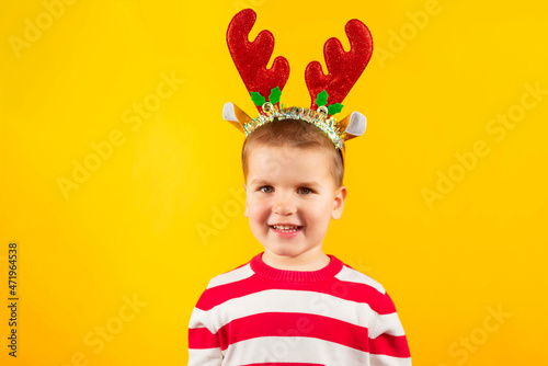 Happy funny toddler boy in horns reindeer with gift on yellow colored background