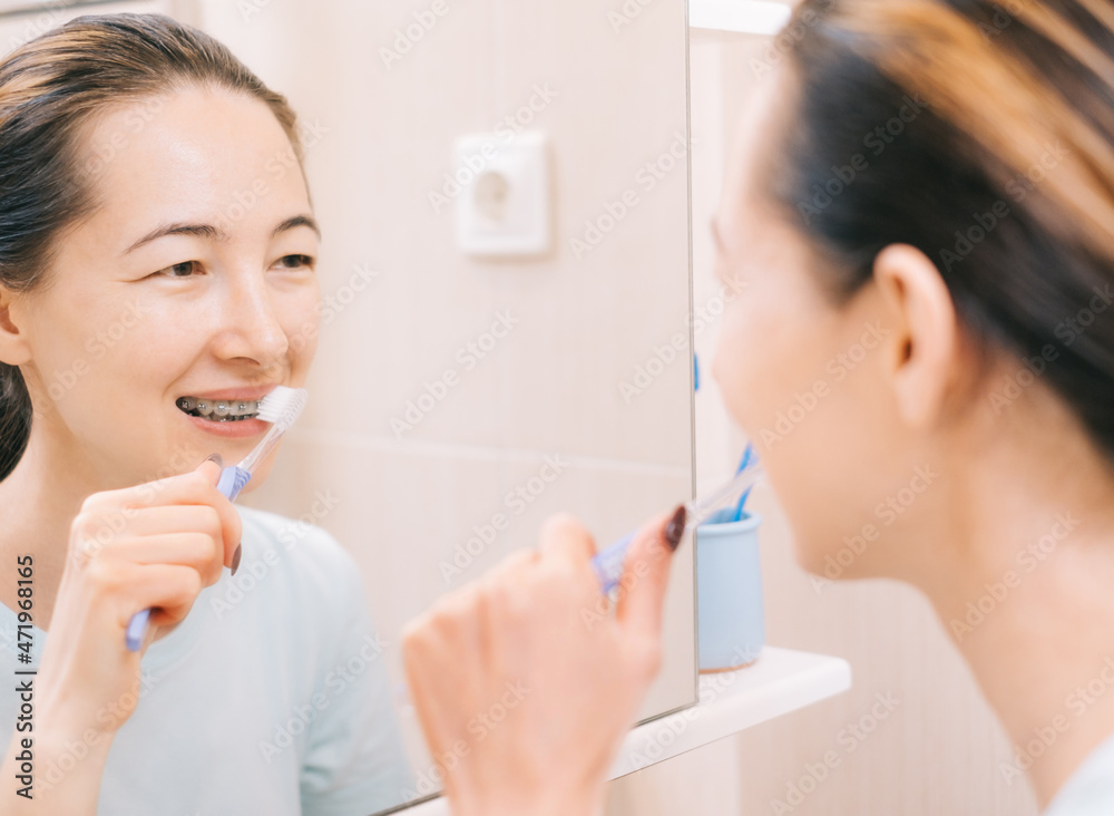 Young woman with braces cleans her teeth with a toothbrush, before mirror. Dental and oral care. Selective focus on the mirror. Young woman with braces cleans her teeth with a toothbrush, before mirror. Dental and oral care. Selective focus on the mirror.