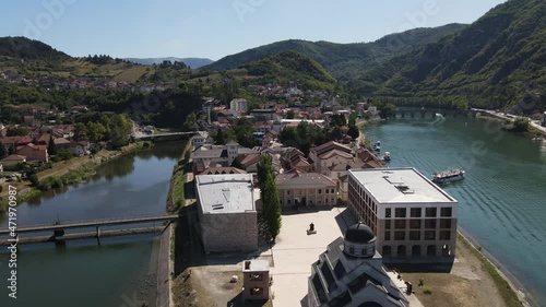 Andricgrad, Visegrad, Bosnia and Herzegovina. Drone Aerial View of Buildings and Church by Drina River With Famous Bridge in Background on Sunny Summer Day photo