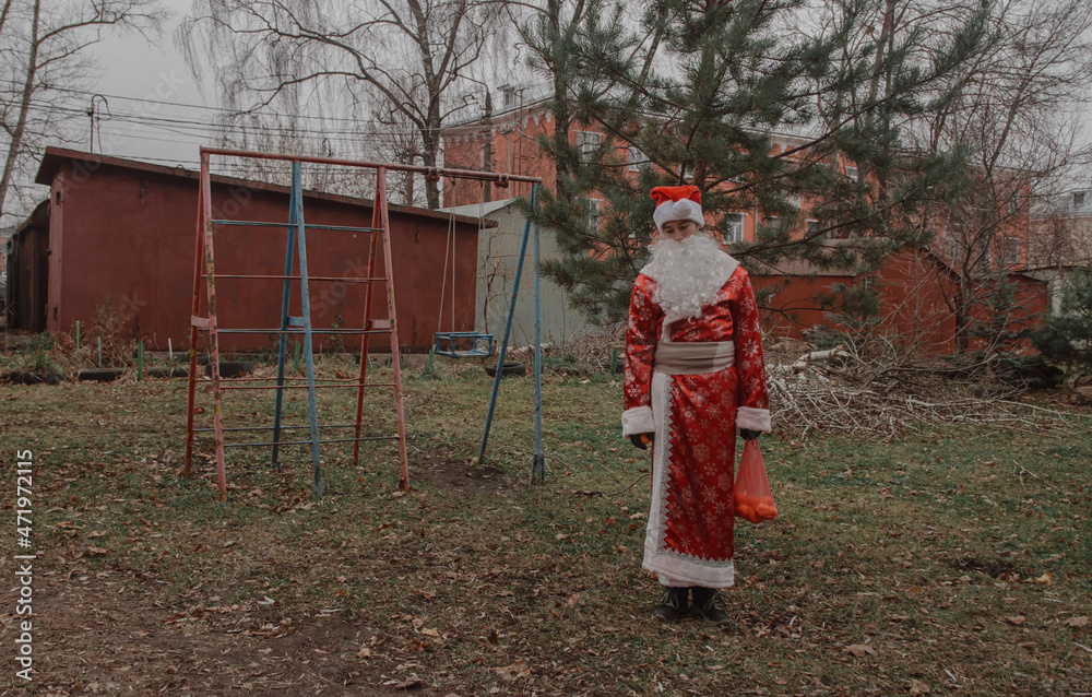 A man dressed as Santa Claus with tangerines in a bag stands on the site next to the garages. The environmental problem of warming when there is no snow in winter. A man dressed as Santa Claus with tangerines in a bag stands on the site next to the garages. The environmental problem of warming when there is no snow in winter.