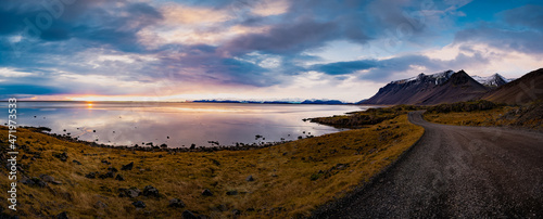 Panoramic sunset photo of road leading along coast of lake to volcanic mountains. High rocky peaks covered with snow layer mirroring on water surface. Driver's point of view on Ring road, Iceland.