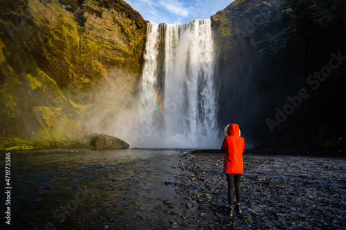 Iceland waterfall travel nature famous tourist destination. Skogafoss waterfall with rainbow and woman under water fall in magical landscape popular Europe attraction.