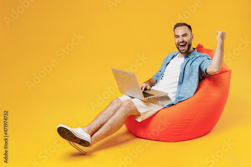 Full body young smiling fun cool happy man wear blue shirt white t-shirt sit in bag chair hold use work on laptop pc computer do winner gesture isolated on plain yellow background studio portrait