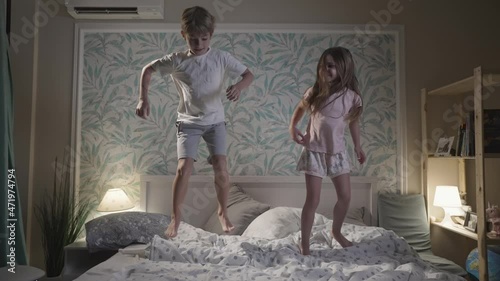 Cute smiling children having fun in bedroom. Happy small kids girl and boy jumping in bed, playing with pillows, enjoying time together at home