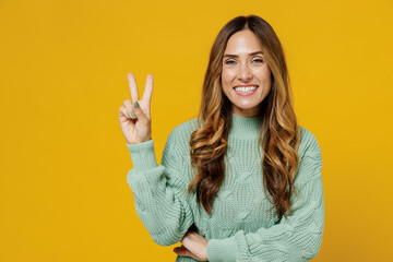 Young friendly cheerful smiling happy cool caucasian woman 30s wearing green knitted sweater showing victory sign isolated on plain yellow color background studio portrait. People lifestyle concept.