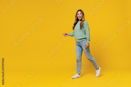 Full body young smiling satisfied happy fun woman 30s wearing green knitted sweater look camera walk go strolling isolated on plain yellow color background studio portrait. People lifestyle concept.