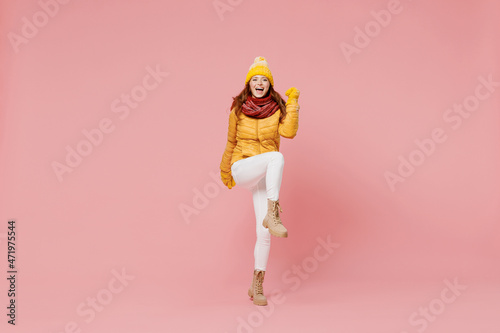 Full size body length cheerful fun young woman 20s wear yellow jacket hat mittens doing winner gesture celebrate clenching fists say yes isolated on plain pastel light pink background studio portrait