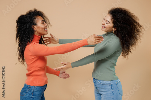 Side view profile two fun young curly black women friends 20s wear casual shirts clothes standing with outstretched hand for greeting hugging isolated on plain pastel beige background studio portrait.