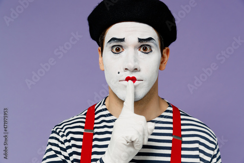 Close up secret vivid young mime man with white face mask wears striped shirt beret say hush be quiet with finger on lips shhh gesture isolated on plain pastel light violet background studio portrait. photo