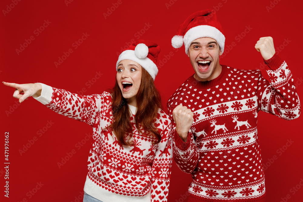Young couple friends two man woman in sweater hat point index finger aside on area do winner gesture isolated on plain red background. Happy New Year 2022 celebration merry ho x-mas holiday concept. Young couple friends two man woman in sweater hat point index finger aside on area do winner gesture isolated on plain red background. Happy New Year 2022 celebration merry ho x-mas holiday concept.