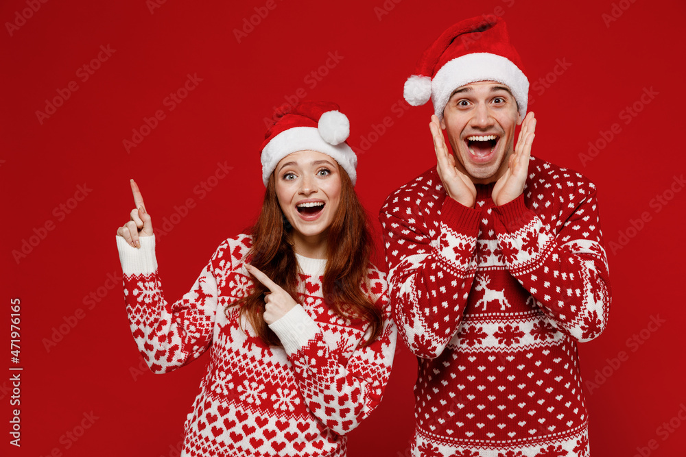 Young fun couple friends two man woman 20s in sweater hat point index finger aside on copy space workspace area holding face isolated on plain red background. Happy New Year 2022 celebration concept. Young fun couple friends two man woman 20s in sweater hat point index finger aside on copy space workspace area holding face isolated on plain red background. Happy New Year 2022 celebration concept.