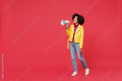 Full size body length fun happy fancy young curly black latin woman 20s wears yellow jacket hold scream in megaphone announces discounts sale Hurry up isolated on plain red background studio portrait.