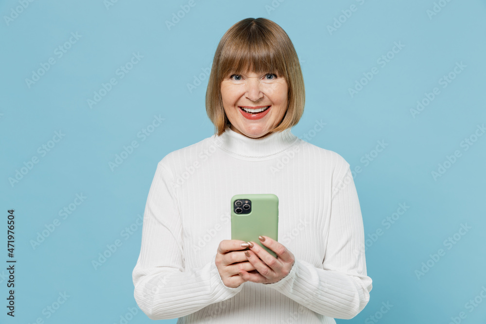 Elderly cheerful woman 50s wear white knitted sweater hold in hand use mobile cell phone browsing internet chatting isolated on plain blue color background studio portrait. People lifestyle concept Elderly cheerful woman 50s wear white knitted sweater hold in hand use mobile cell phone browsing internet chatting isolated on plain blue color background studio portrait. People lifestyle concept