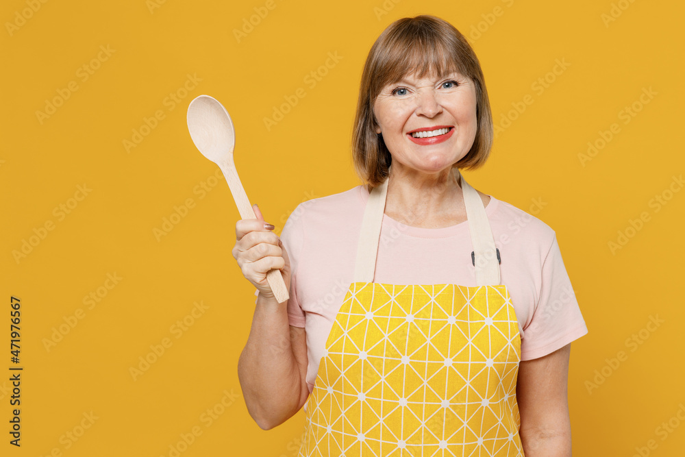 Elderly fun cheerful happy cooker housekeeper housewife woman 50s in orange apron hold empty spoon try meal eat isolated plain on yellow background studio portrait. People household lifestyle concept.