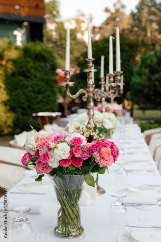 a bouquet of white roses  pink roses  pink carnations on a festive table with dishes decorated with large candelabra on a background of greenery of garden