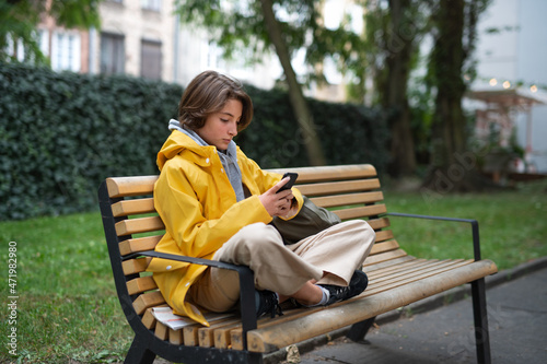 Preteen schoolgirl sitting on bench and using smartphone outdoors in town.