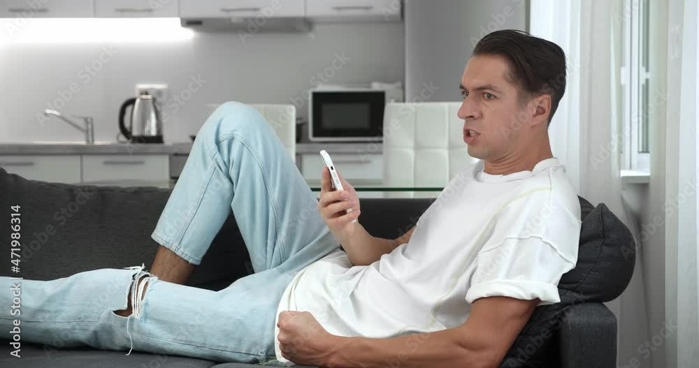Annoyed young man holding smartphone frustrated by bad message, resting on sofa at home. Angry male uses application on his mobile phone, bangs his fist on the sofa and shouts. Display of aggression.