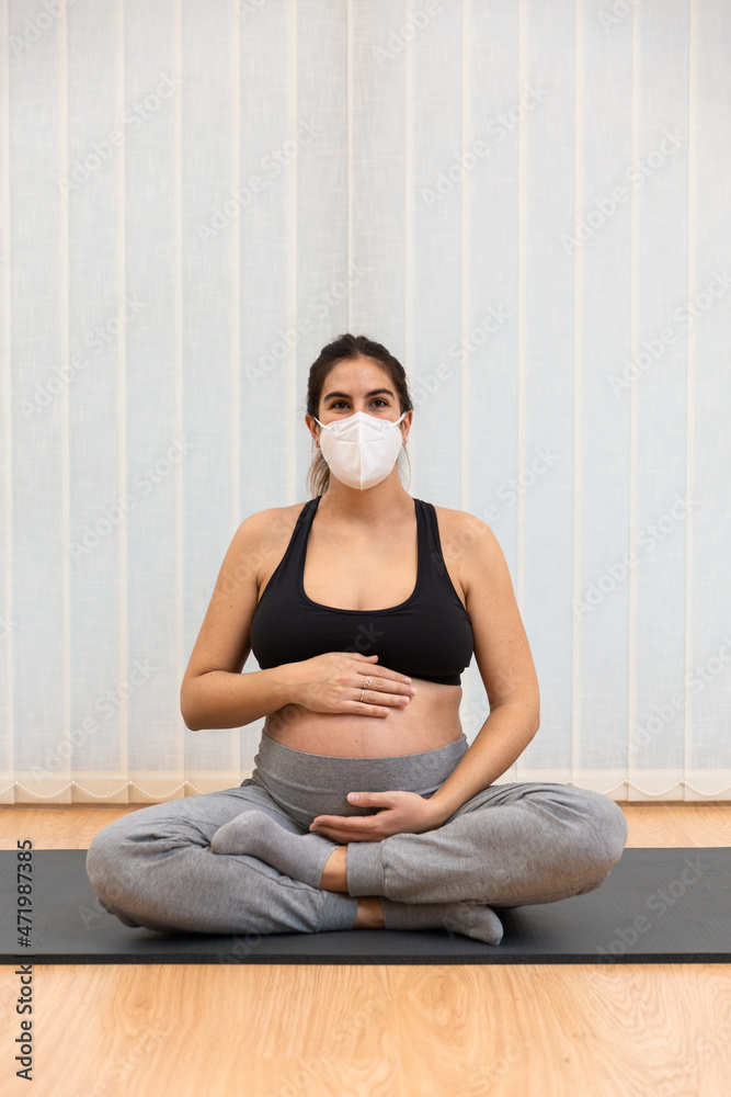 A healthy pregnant woman with face mask sitting on a mat doing yoga at home
