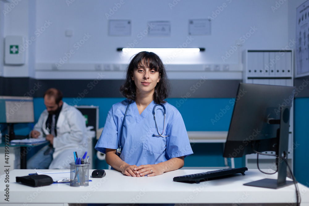 Portrait of medical assistant using computer at desk for work overtime. Woman nurse in uniform looking at camera preparing for paperwork and healthcare checkup, working late at office. Portrait of medical assistant using computer at desk for work overtime. Woman nurse in uniform looking at camera preparing for paperwork and healthcare checkup, working late at office.
