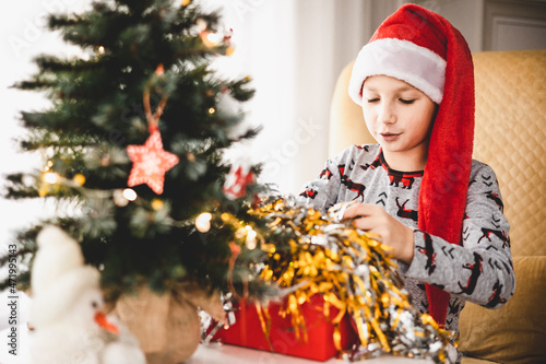 Christmas and New year vacation. Happy child boy in pajamas and red Santa hat opening gift box in early morning at home. Winter holiday spirit