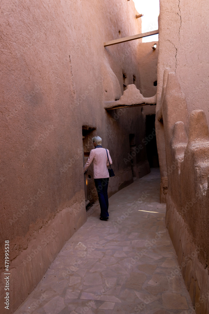 A lonely Western female tourist standing in the narrow street in Ushaiqer Heritage Village, Saudi Arabia. Back view. A lonely Western female tourist standing in the narrow street in Ushaiqer Heritage Village, Saudi Arabia. Back view.